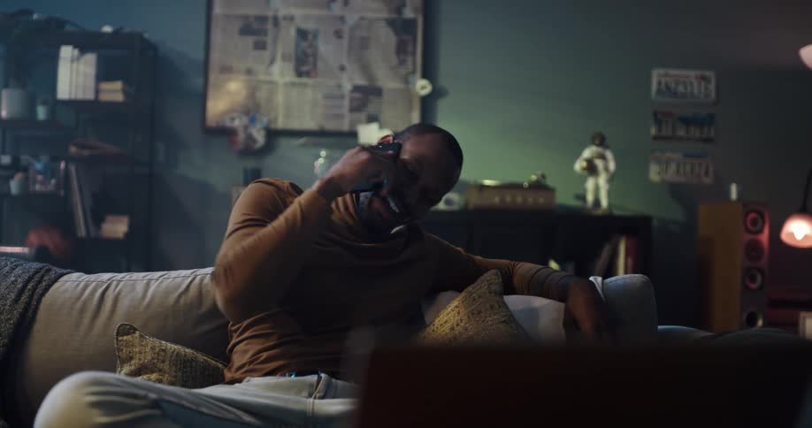 Happy young man sitting on a couch in the living room at home talking on the phone