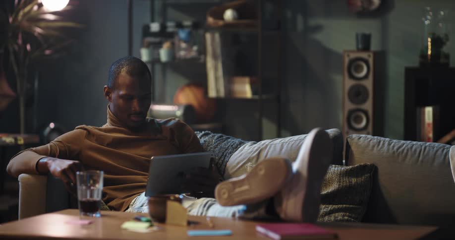 Man sitting down on the couch of his living room putting his legs up in the center table and using his ipad