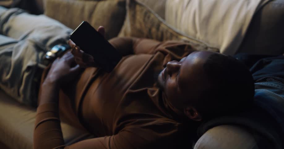 A black man is lying on the couch holding his phone and talking