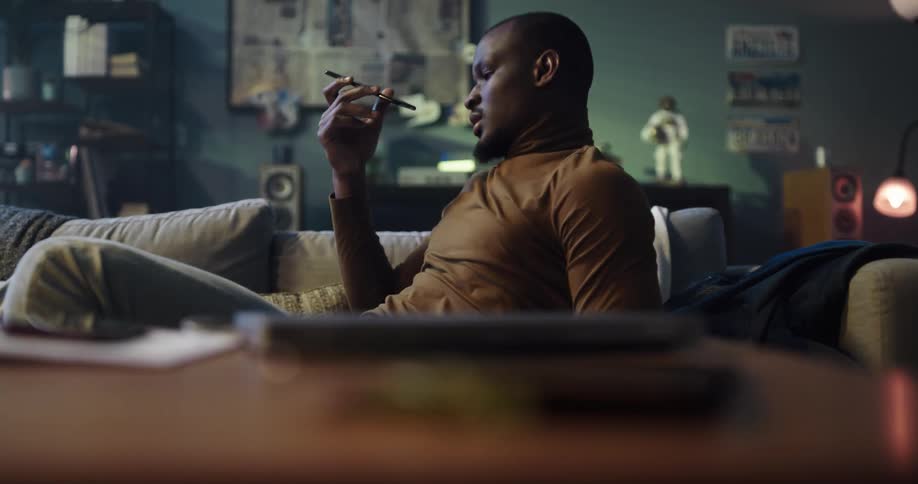 Man with brown turtleneck sweater and jeans lies on couch and answers call on smartphone in living room at home