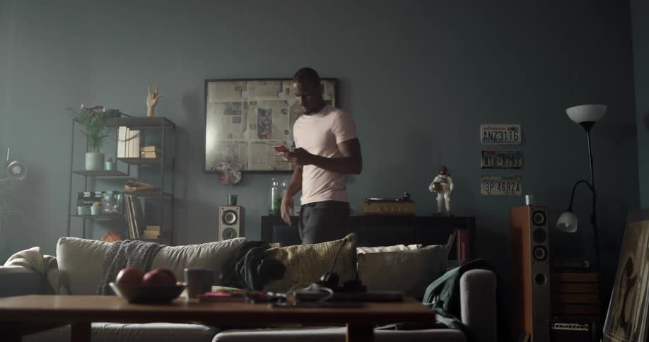 Man with white t shirt and jeans walks in living room using smartphone