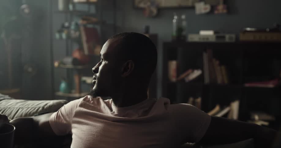 Black man sitting on the couch drinking a cup of coffee and then grabing his phone from the table in front of him