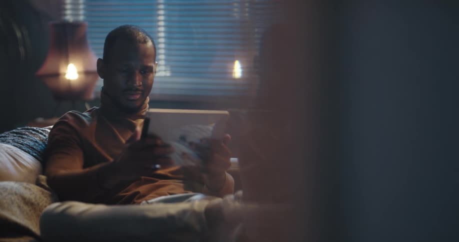 Man with brown turtleneck sweater and jeans sits on couch using tablet and smiling in living room at home