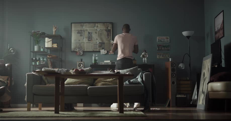 Man sits on a sofa and looks at a record album then gets up and walks to a record player and puts the album on it