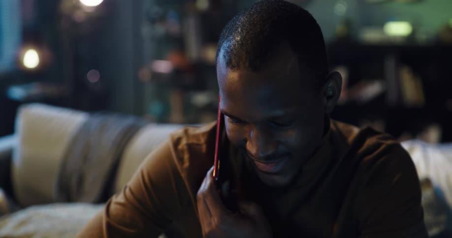 Man with brown turtleneck sweater sits on couch talking on smartphone in living room at home