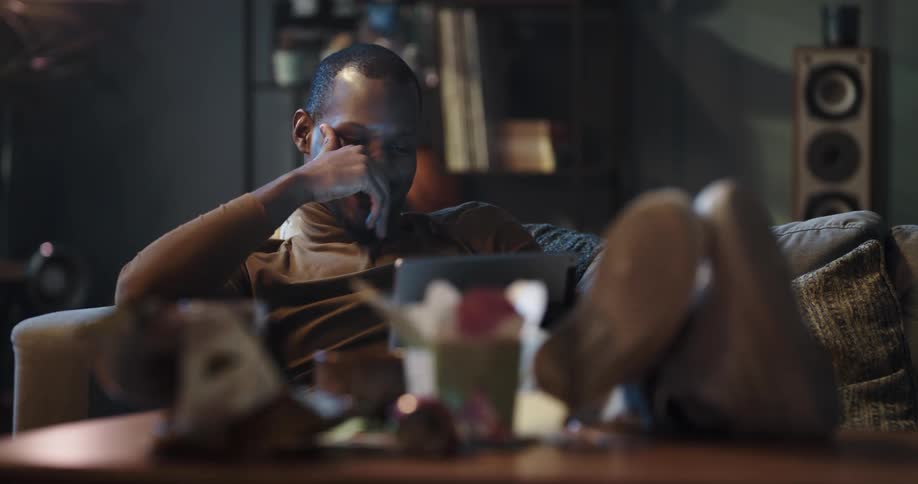 Man relaxing on the couch in the living room at home smiles and laughs looking at a tablet