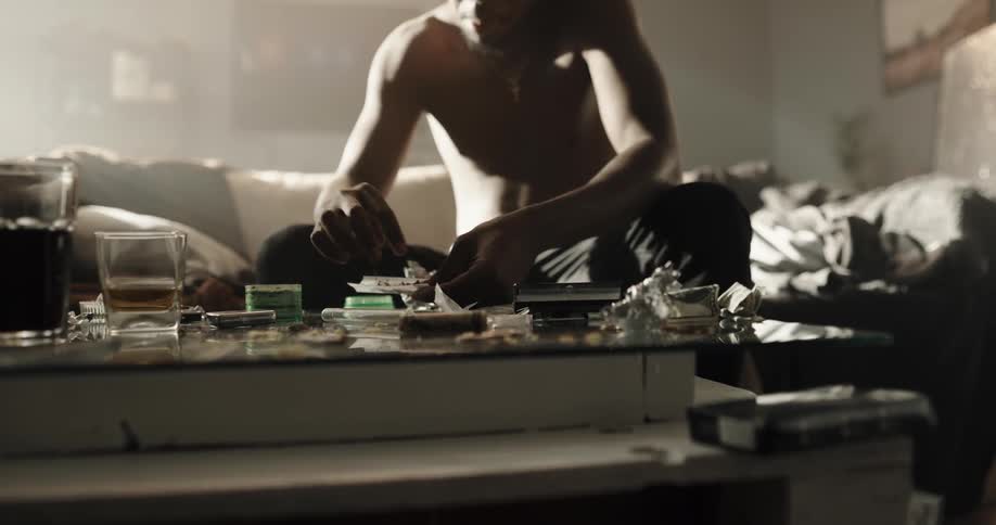 Drug addict sitting on a couch at home fills a handmade cigarette over a coffee table filled with drug paraphernalia