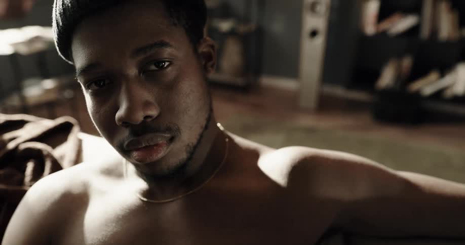 Shirtless black man wearing a toque raises his head as he sits on a sofa