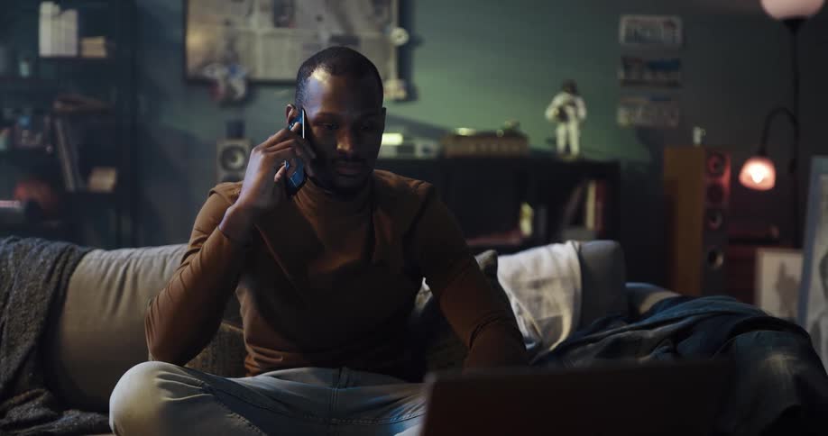 A black man is sitting on his couch talking on the phone