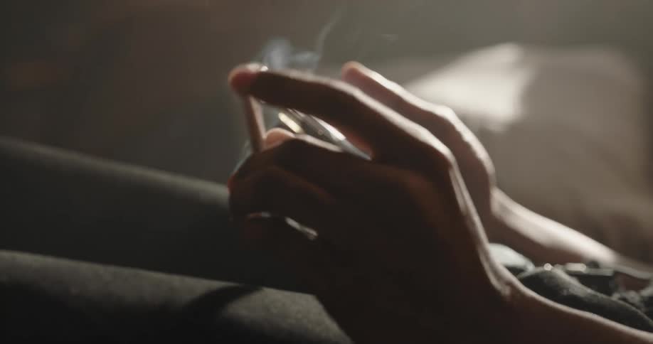 A man is holding his cell phone and looking at it as he plays with a lit cigar in his other hand and smokes