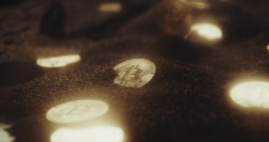 Close up of the shiny engraved surface of golden bitcoin physical coins over dark sand blowing in the wind