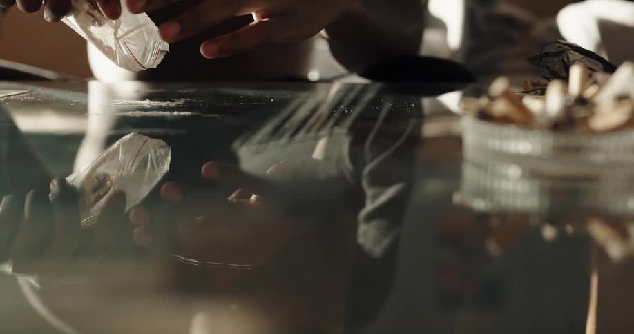 Table surface reflects barechested man as he empties powder onto surface near a drink a credit card and a full ashtray