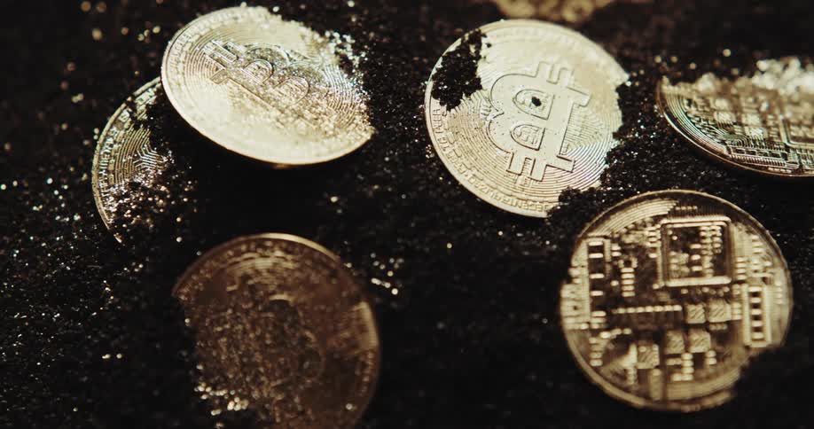 A number of shiny coins with the bitcoin logo on them are partially buried in black powder