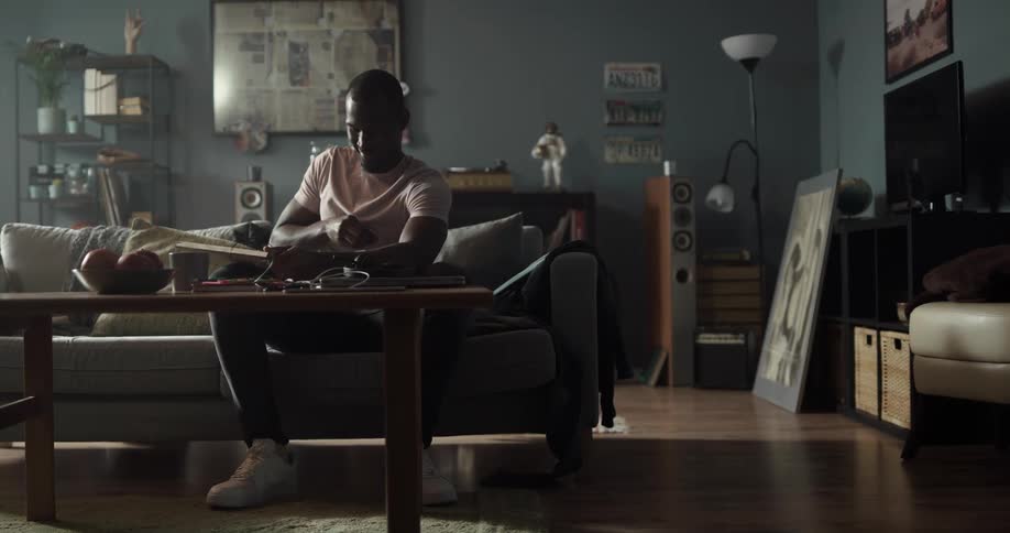 A man carries a package into his living room sits on the couch and smiles as he opens it