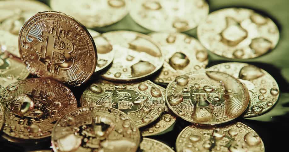 A number of shiny coins with the bitcoin logo on them are piled on a wet surface