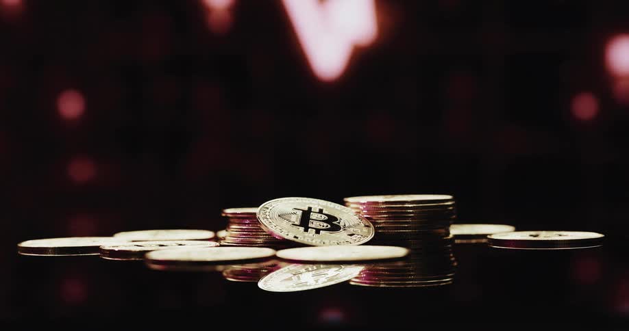 Stack of golden coins with the bitcoin logo printed on them in front of a screen showing a financial graph