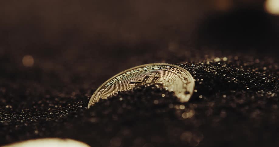 Close up of the shiny engraved surface of golden bitcoin physical coins over dark sand blowing in the wind
