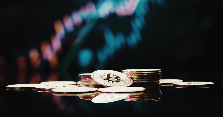 Stack of golden coins with the bitcoin logo printed on them in front of a screen showing a financial line graph