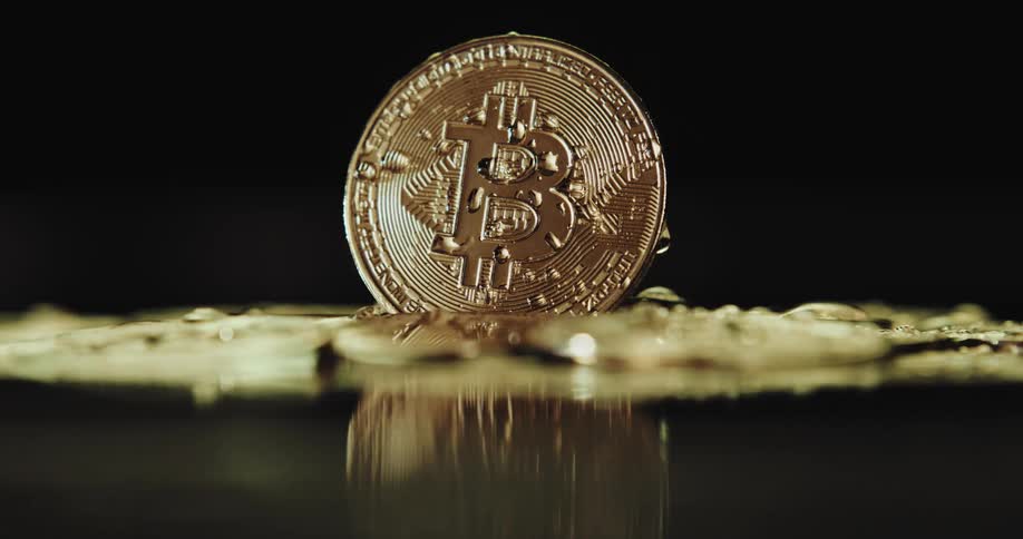 A single shiny coin with the bitcoin logo on it stands atop a wet surface