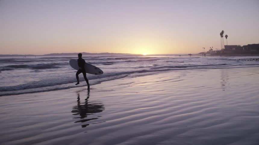 A man is running towards the water with a surfboard at sunset