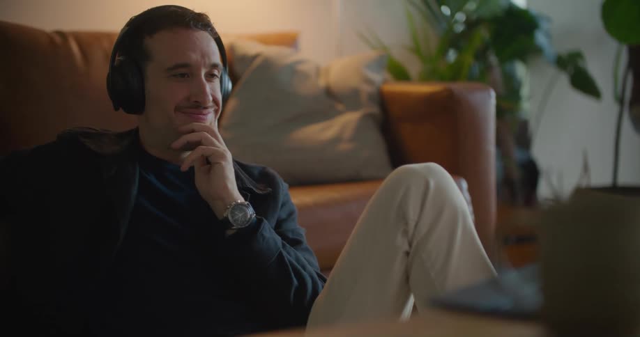 White man sitting on the floor in his living room wearing a headphone and smiling while looking at something on his laptop at the center table