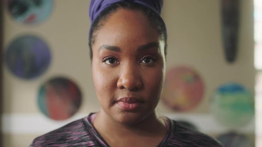 A black woman calmly looks ahead at the camera and blinks twice