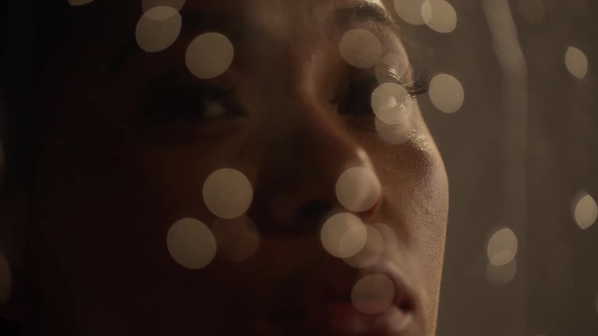 A woman turns to look at the camera while water droplets roll down a piece of glass in front of her