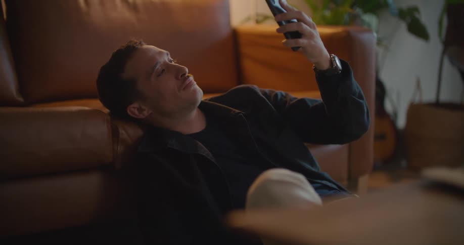 Man sitting on the floor leaning his head against the couch looking at the ceiling and then and looking at his phone