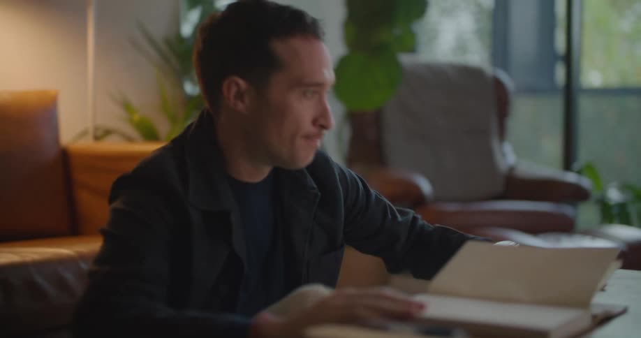 Man leans against the sofa as he thinks then sits up at a table with a laptop on it then lowers his head on a book in frustration
