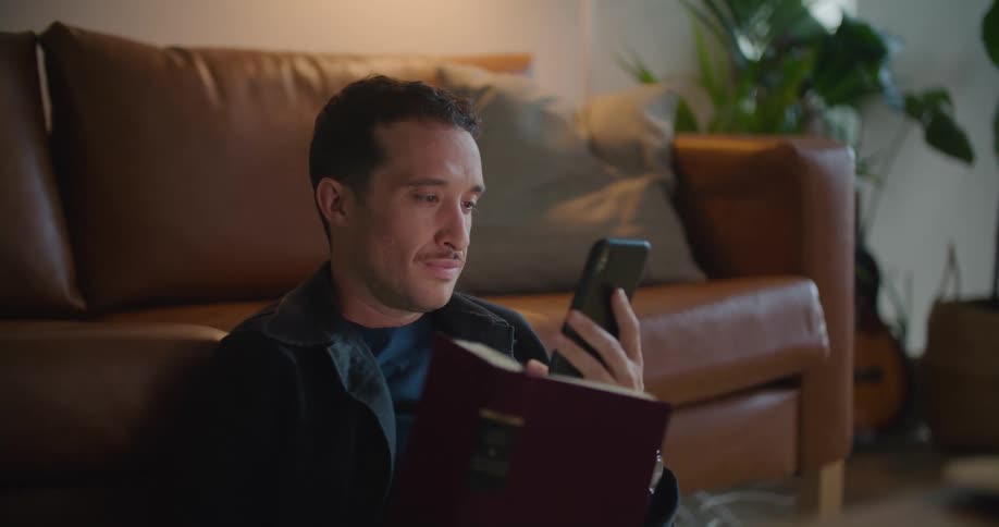 White man sitting on the floor in his living room reading a book and then using his smartphone