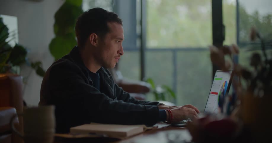 A man sitting at a desk looks over at his notebook while typing on his laptop computer