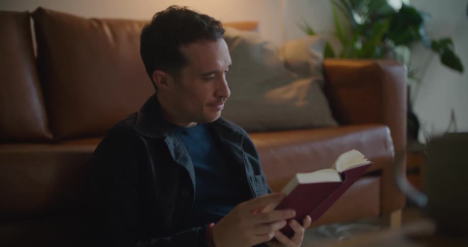 Man with naval blue jacket shirt brown pants reads book sitting on floor leaning against brown couch in living room