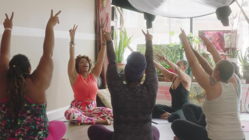 A group of women are seated in a circle on the floor while stretching their arms above their heads
