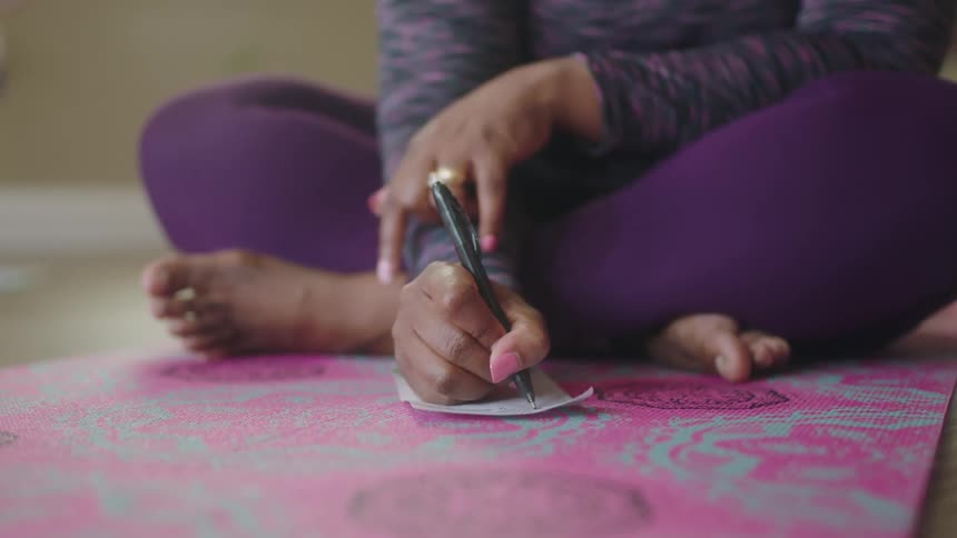 A woman sitting on a yoga mat writes on a small piece of paper in front of her