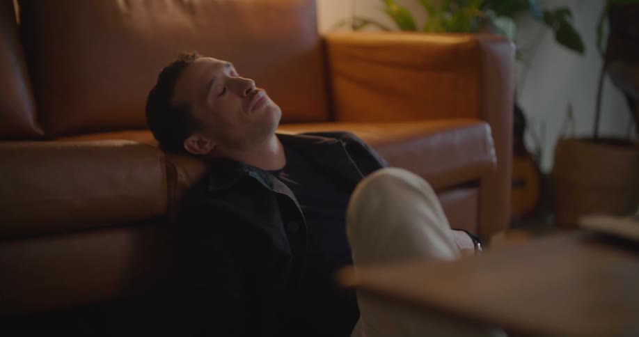 Thoughtful man sitting on the floor with his head leaning against the couch behind him