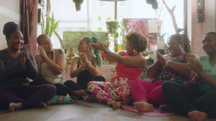 A woman leads others in clapping joyfully while they sit on mats on the studio floor