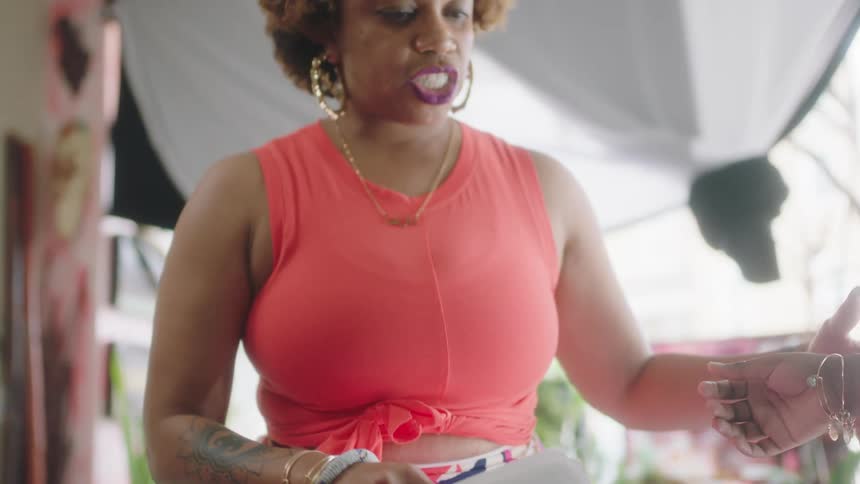 Woman wearing hoop earrings and holding papers in her hand bends over and talks to another woman while a hand reaches out next to her