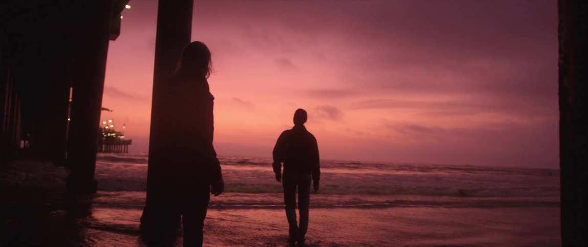Man and woman silhouetted against the red sky move among pier supports on the beach
