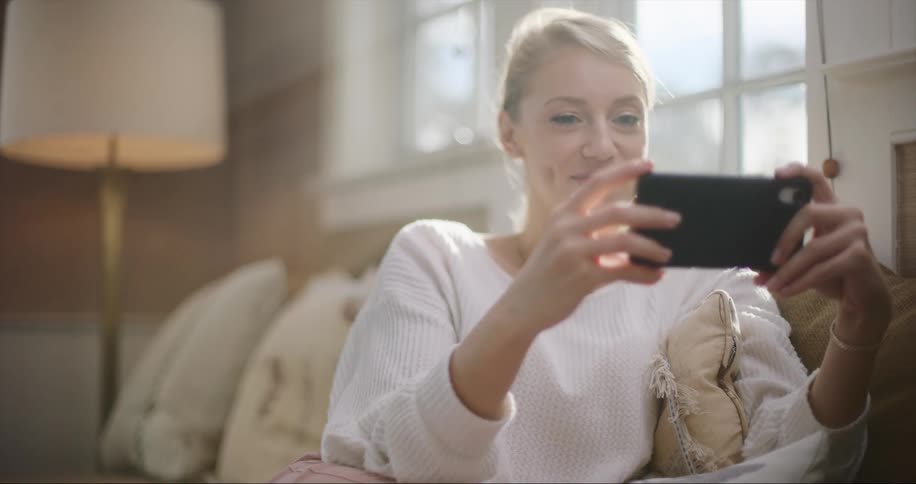 Blonde woman sitting on couch rotates cell phone to landscape before viewing screen with a smile
