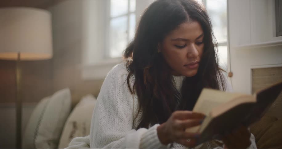 Woman with dark wavy hair and white sweater reads book in living room at home