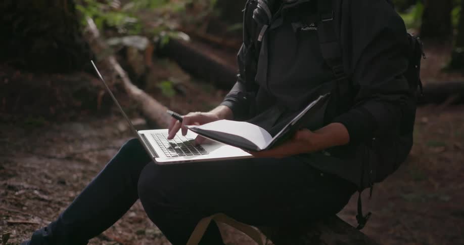 A man in glasses types on a laptop in the woods