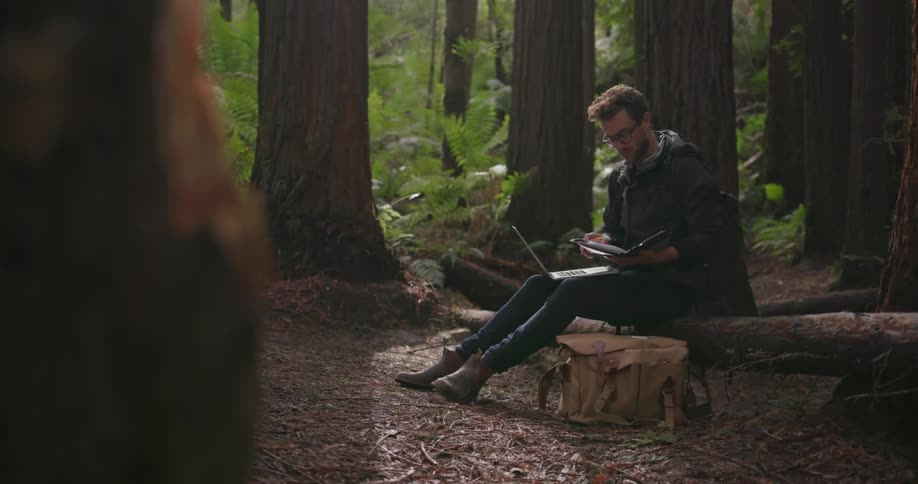 A man is sitting on a log in the woods looking at his book and laptop
