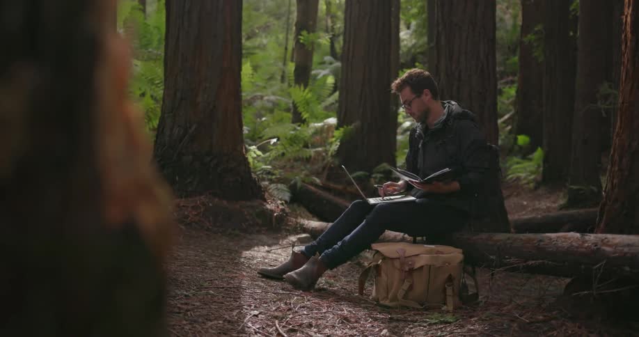 A man is using his laptop and notebook while sitting in the forest