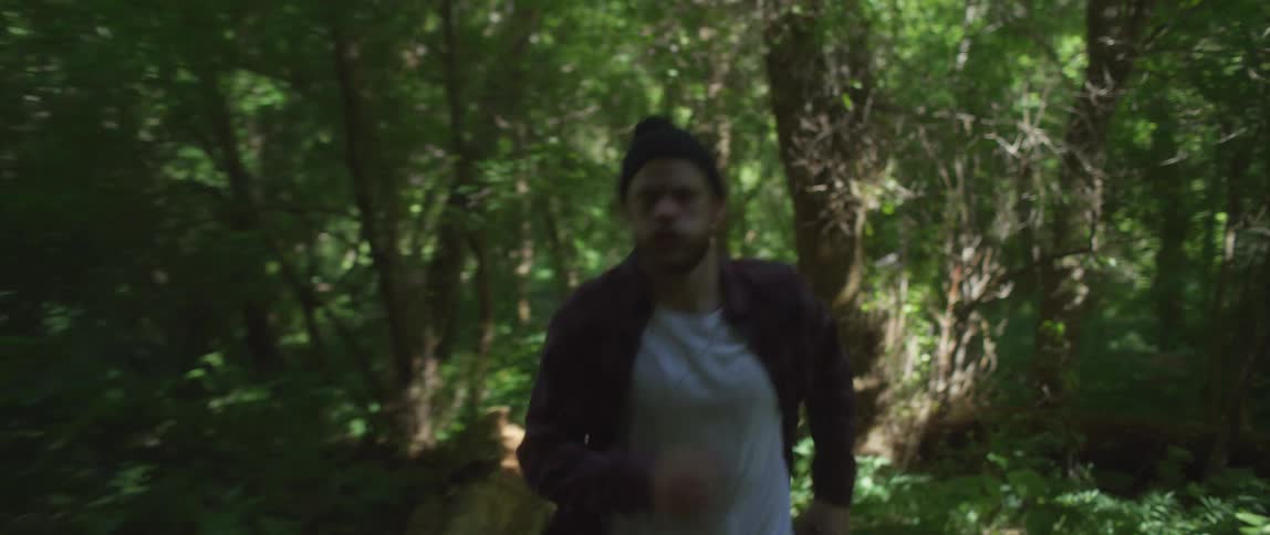 A man wearing a flannel shirt and a knit cap is running on a trail in the woods