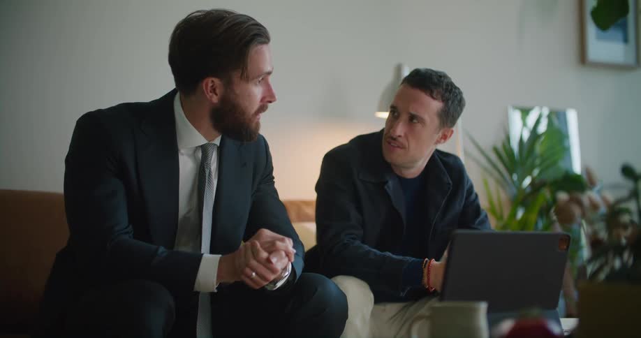 Business man sitting on the couch talking to another man next to him and looking at a laptop on the center table in front of him