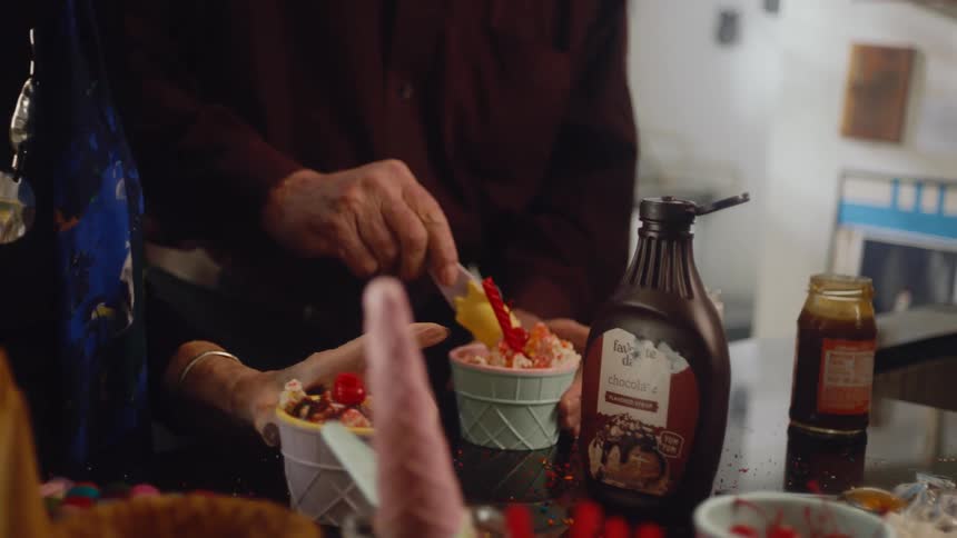 An elderly couple prepare small bowls of ice cream and eat them together while standing in a home kitchen during the day