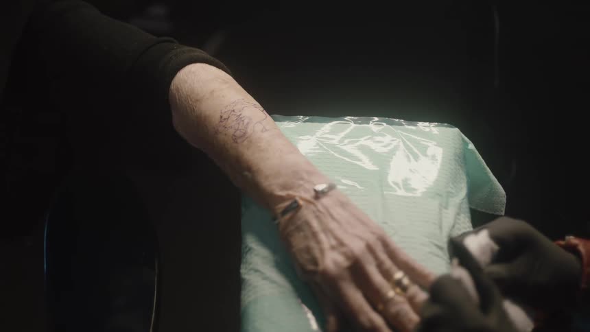 Young female tattoo artist wearing black latex gloves drawing on an elderly womans forearm