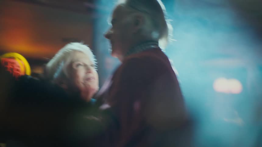 Elderly man with red sweatshirt and elderly woman with short gray hair dace in spotlight in room