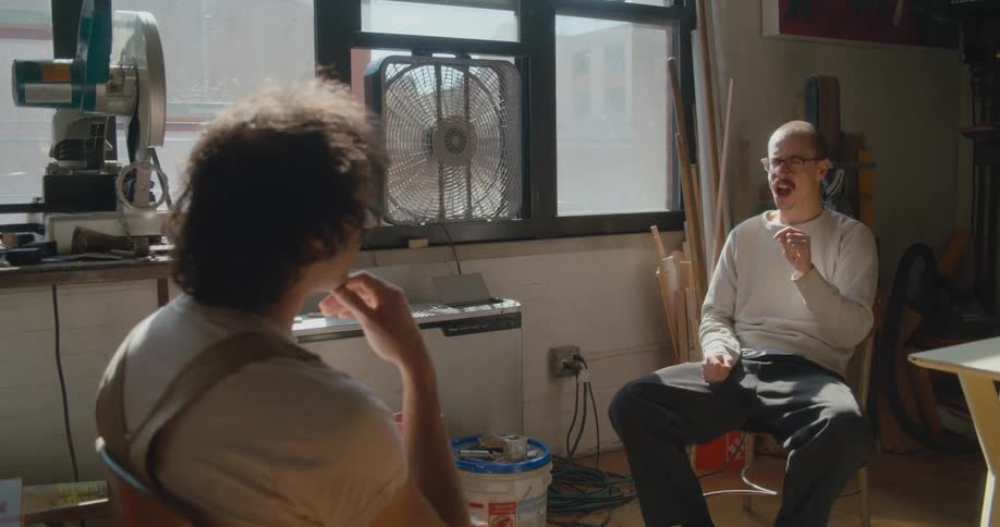 Two men sitting face to face talk to each other with sign language next to a window with fan in painting studio