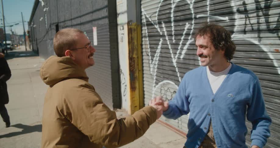 Two men are standing on a sidewalk talking in sign language and shake hands as one man pats the other on the back